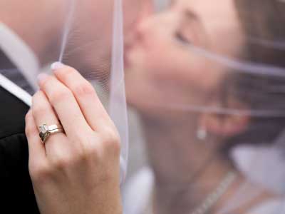 A Newly Married Couple Kissing Under The Bride's Veil With The Focus On Her Wedding Ring